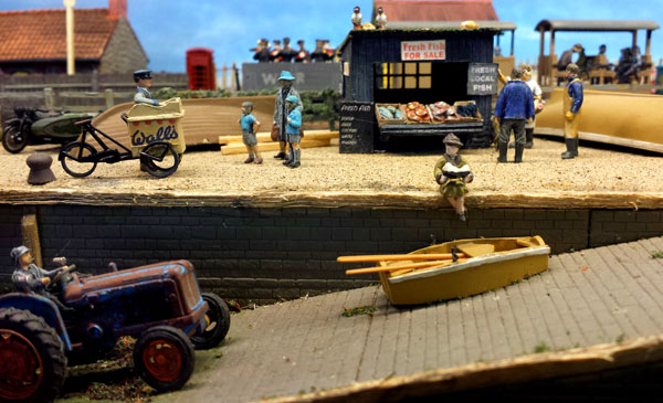 model of 1950s seaside. woman sits reading a book while two boys queue for ice cream