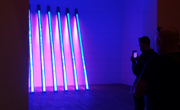 two men in a dark room photograph some neon artwork on a phone. pink and purple colours
